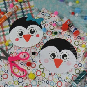 A colorful penguin craft setup featuring two round penguin cards made from a free printable, surrounded by craft supplies like scissors, glue, markers, and pom-poms on patterned paper.