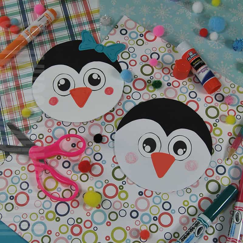 A colorful penguin craft setup featuring two round penguin cards made from a free printable, surrounded by craft supplies like scissors, glue, markers, and pom-poms on patterned paper.