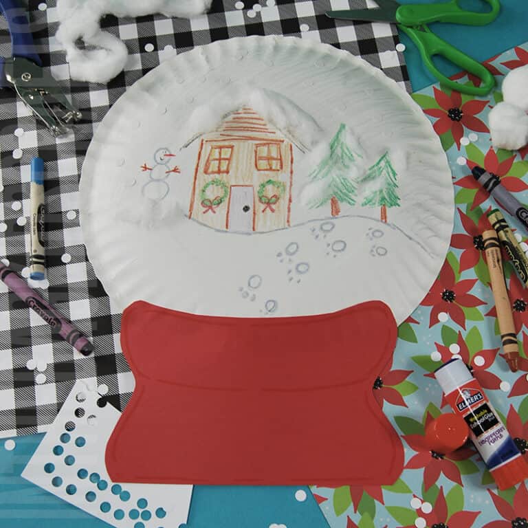A handmade paper plate snow globe craft featuring a winter scene with trees and a house, designed as a fun preschool craft that doubles as a fine motor activity and creative winter craft for kids.