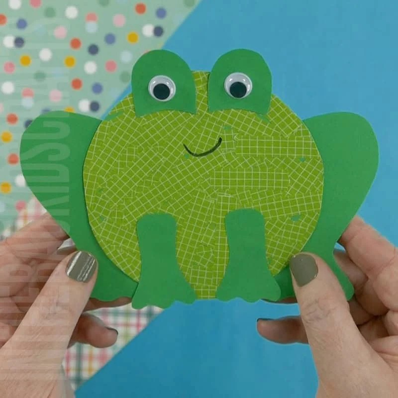 A preschool frog craft made from green paper with googly eyes, held by a hand against a colorful background, designed as a fine motor activity for young kids.