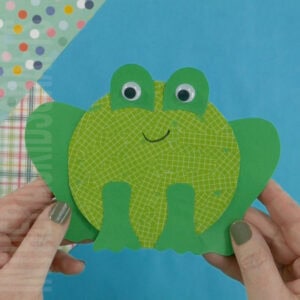 A preschool frog craft made from green paper with googly eyes, held by a hand against a colorful background, designed as a fine motor activity for young kids.