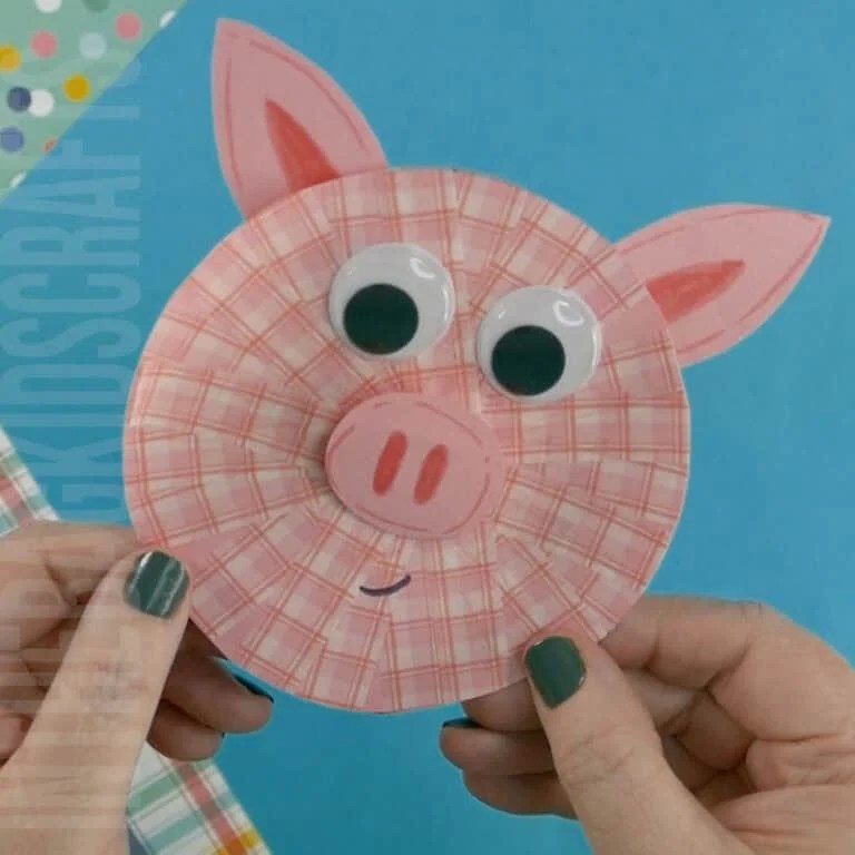 A pair of hands holds a completed pig craft, made from cut-out paper pieces, as part of a preschool craft and fine motor activity against a colorful background.A pair of hands holds a completed pig craft, made from cut-out paper pieces, as part of a preschool craft and fine motor activity against a colorful background.