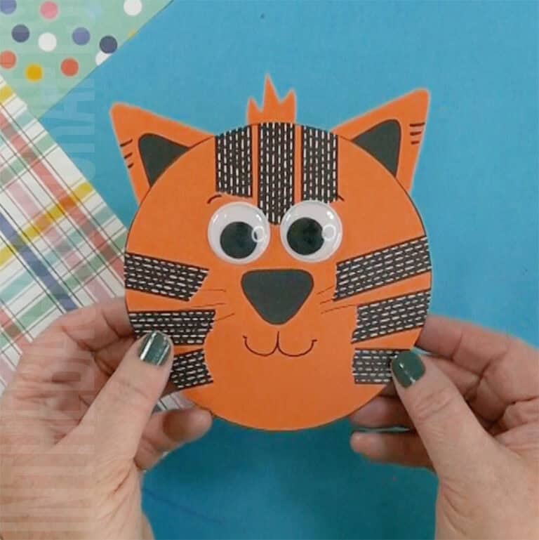 A child’s hands hold a round orange tiger made from paper and craft supplies as part of a preschool tiger craft and fine motor activity.