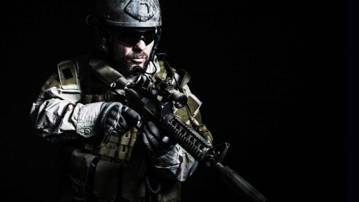 Military soldier in tactical gear with rifle in dark background.