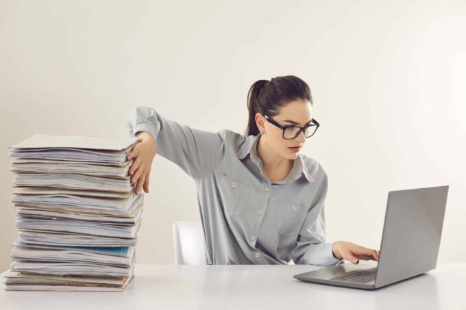 Overwhelmed woman managing paperwork and digital tasks at her desk.