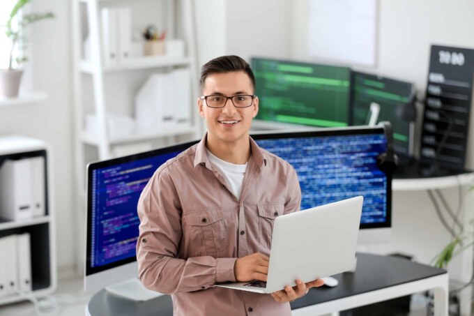 IT professional working on a laptop with multiple monitors displaying code and IT management tools.
