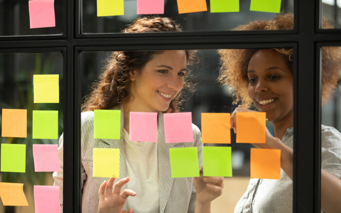 Two women collaborating with sticky notes on glass wall for agile project management.