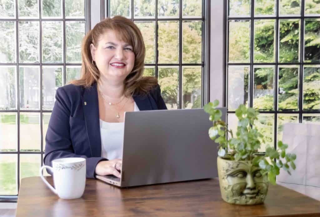 Jennifer typing on laptop with plant and mug on desk and windows in background