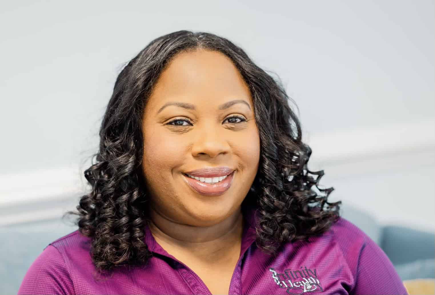 A woman with curly hair smiling at the camera, wearing a purple shirt with a logo on it.