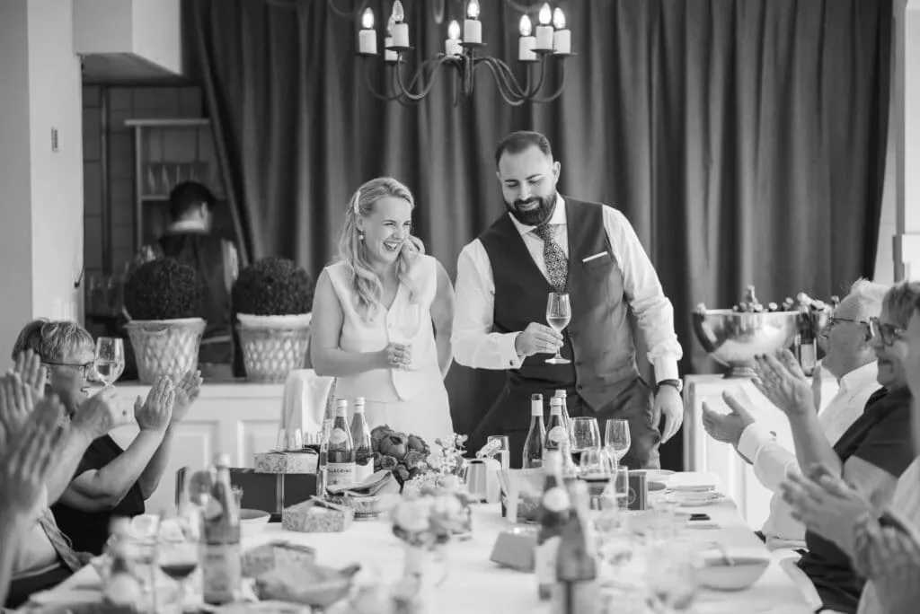 Hochzeitsfotografie mit Herz: Glückliches Brautpaar bei Feier mit Freunden, lachend und anstoßend.