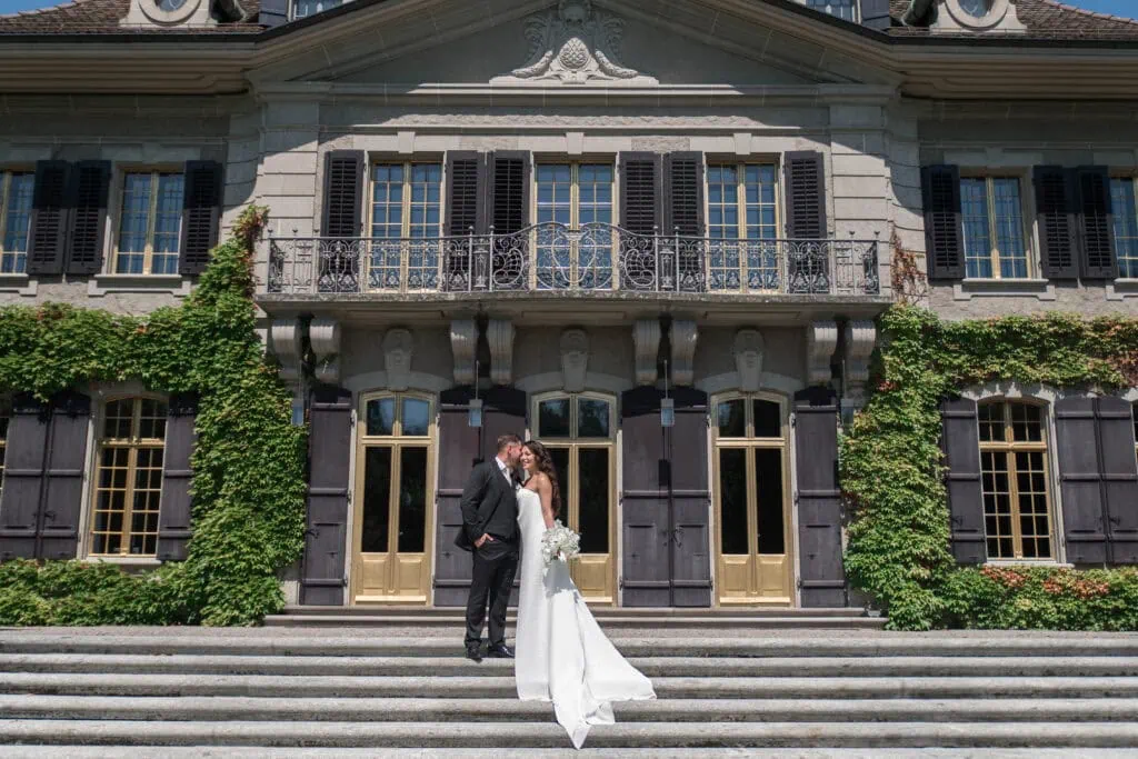 Hochzeitspaar vor einem alten, beeindruckenden Gebäude mit Treppen und grünen Pflanzen.