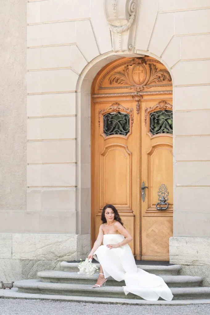 Braut in weißem Kleid sitzt vor einer kunstvollen Holztür bei Hochzeitsfotos.