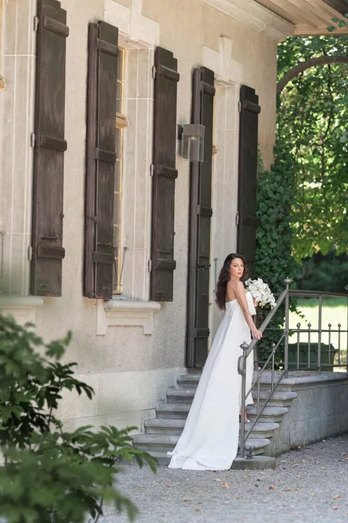 Braut in weißem Hochzeitskleid auf Treppe vor altem Gebäude mit Holzfenstern.