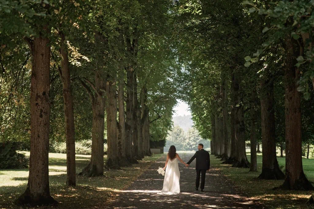 Hochzeitspaar spaziert Hand in Hand durch einen Baumweg im Park.