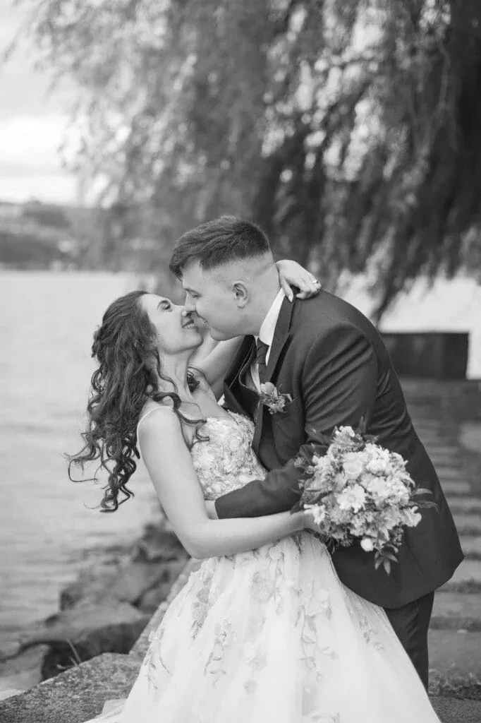 Hochzeitspaar küsst sich am Flussufer, romantische Stimmung, Naturhintergrund.