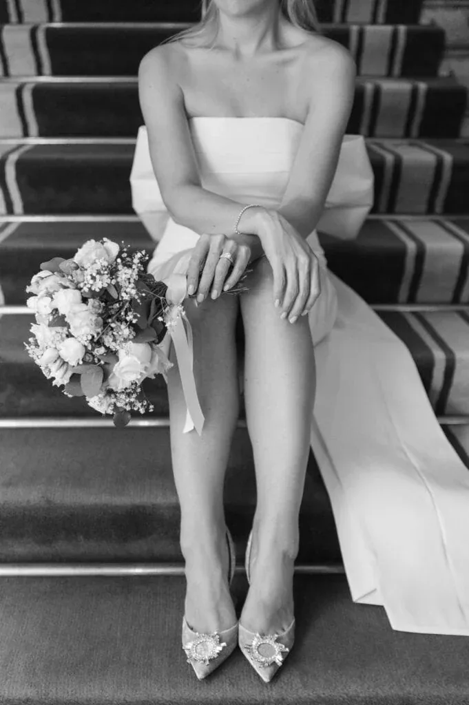 Braut in weißem Kleid mit Blumenstrauß auf Treppe sitzend, schwarz-weiß.