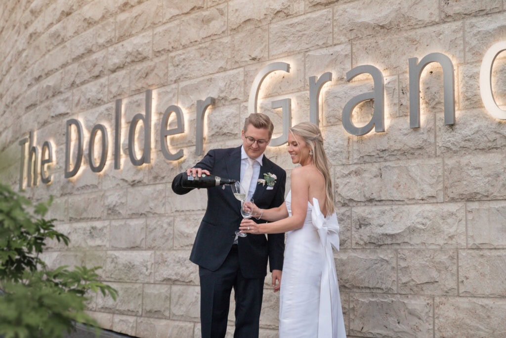 Hochzeitspaar beim Anstoßen mit Sekt vor einer Steinmauer mit Schriftzug.