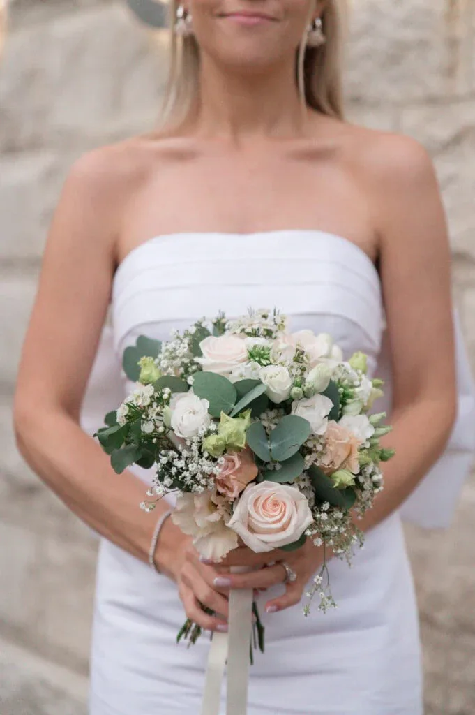 Hochzeitsstrauß mit Rosen, Eukalyptus und weißen Blumen, gehalten von einer Braut in weißem Kleid.