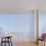 Spacious living space with large windows featuring vertical blinds, minimalist furniture, cozy armchair, and decorative accents, perfect for stylish, natural light-filled interiors.