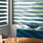 Bright room with layered blue shutters, sunlight filtering in, and a stylish glass bowl on a teal cabinet.