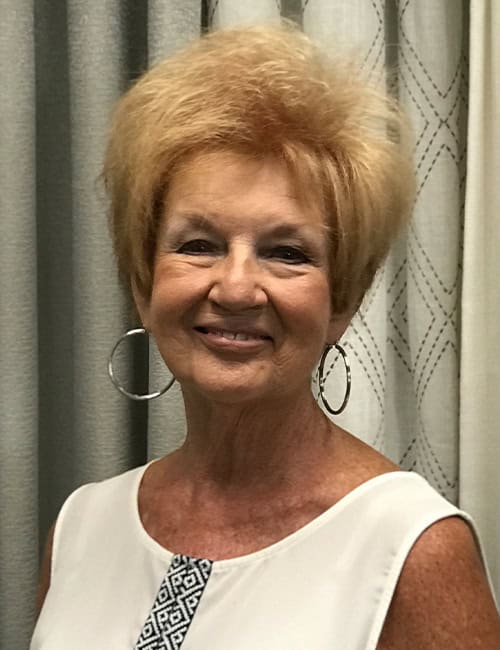 Elegant elderly woman smiling, wearing a white sleeveless top with a decorative neckline, large hoop earrings, standing in front of patterned curtains.