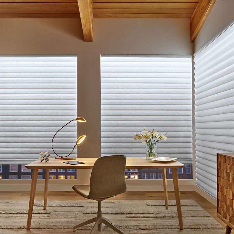 Bright workspace with blinds, wooden desk, and comfortable chair. Elegant lamp, flower vase, and city view create a peaceful work environment. Perfect for home or office use.