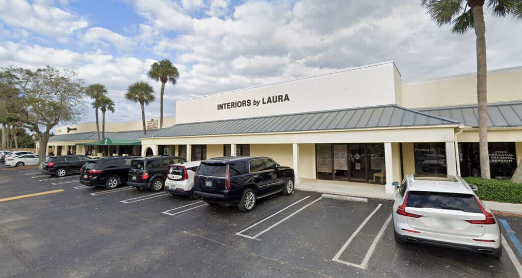 Modern retail storefront image of In The Shade Florid, showcasing the exterior of a gentle commercial building with a parking lot, palm trees, and a partly cloudy sky, emphasizing shopping and interior design services.
