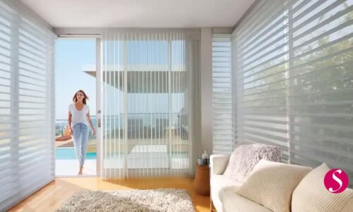 Smooth white horizontal blinds in a modern living room with a woman enjoying sunlight and outdoor views, showcasing sunshade window treatments by In The Shade for stylish, functional window coverings.