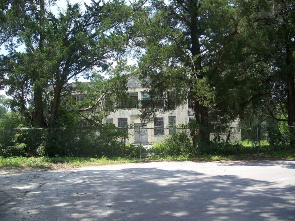 Overgrown abandoned building behind a wire fence, surrounded by large trees with extensive foliage, casting shadows onto the dirt road in the foreground, creating a scene of urban decay and nature reclaiming space.