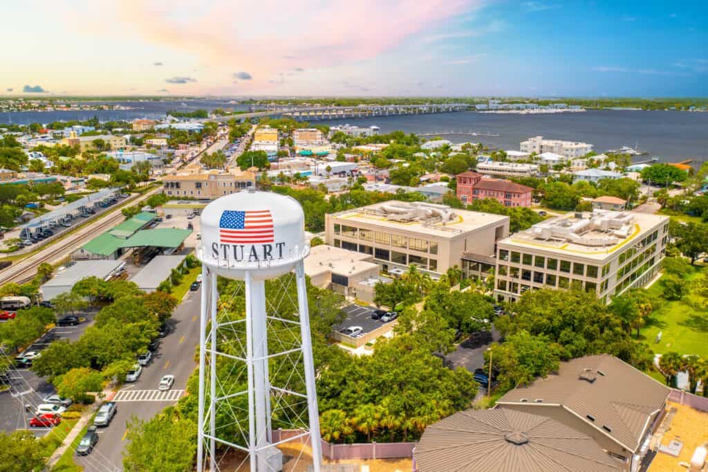 Colorful aerial view of Stuart, Florida skyline with a prominent water tower, lush greenery, and waterfront buildings along the St. Lucie River, showcasing the scenic coastal community and vibrant downtown area.