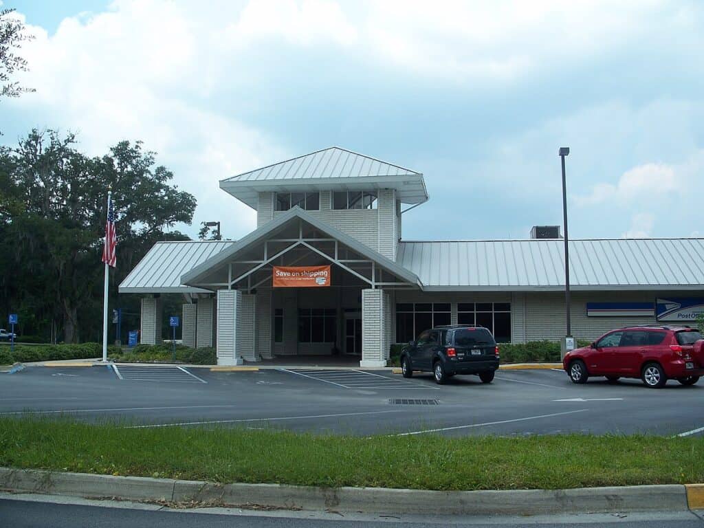 Post Office building with parking lot and American flag, modern design, efficient service, accessible entrance, eco-friendly infrastructure, USPS facility, shipping services, community hub, postal services, urban architecture.