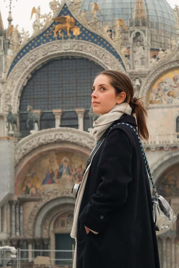 A woman in a dark coat and scarf stands in front of St. Mark’s Basilica in Venice, Italy. The intricate domes and golden mosaics of the historic landmark create a stunning backdrop