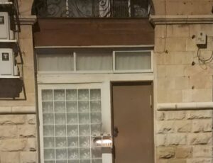 דלת ירושלמית סגורה, לצידה תיבת נרות מוארת – רמז לריח חמים שמתחיל בפנים