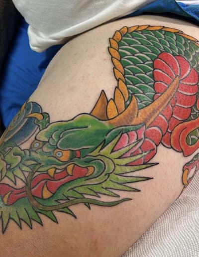 Jesse Britten Tattoo, St. Augustine, FL Dragon tattoo on a thigh, featuring green, red, and gold colors.