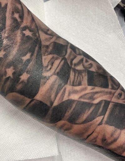 Jesse Britten Tattoo, St. Augustine, FL Black and grey forearm tattoo of a waving American flag with stars and stripes.