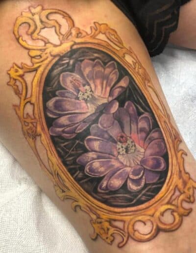 Jesse Britten Tattoo, St. Augustine, FL Traditional tattoo of purple flowers in an ornate gold frame on a thigh.
