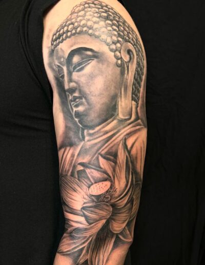 Buddha head tattoo with lotus flower on arm, detailed black and gray ink.
