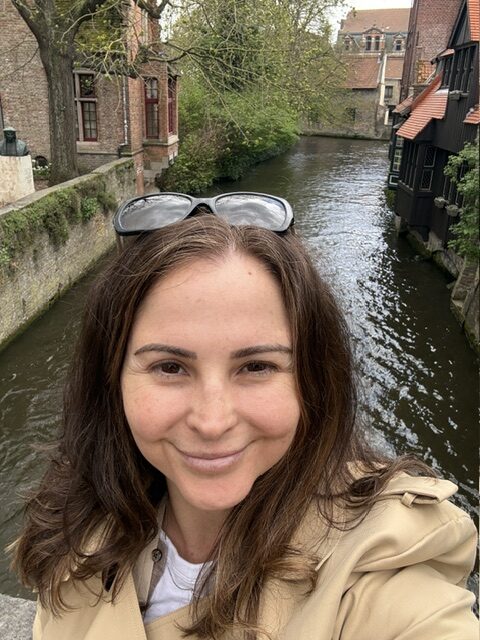 A woman taking a selfie by a canal in Bruges, Belgium, with historic buildings and lush greenery in the background.