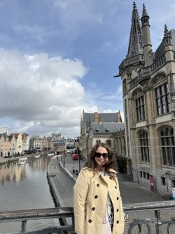 A woman in sunglasses and a beige coat standing on a bridge overlooking a historic city with a river and medieval buildings.