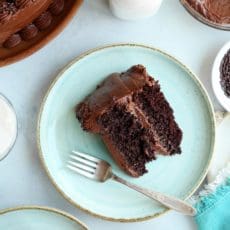 Slice of chocolate cake on a pale green plate with a fork and chocolate frosting.