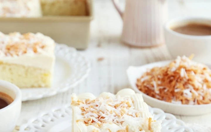 Slice of coconut cake on small white plate with coffee.