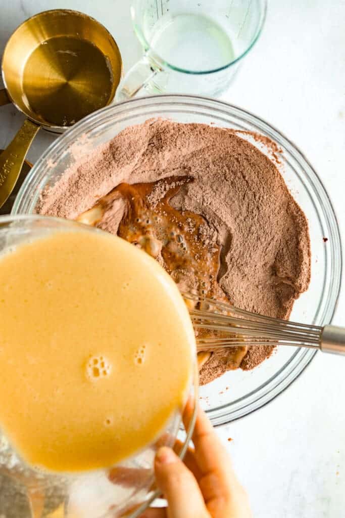 A bowl of beaten eggs being added to a second bowl of dry ingredients with gold and glass measuring cups.