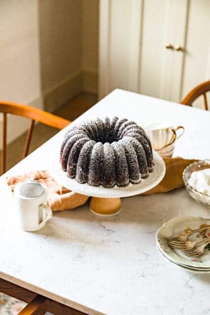 A Chocolate Bundt Cake on a Ceramic cake plate on top of a Marble countertop