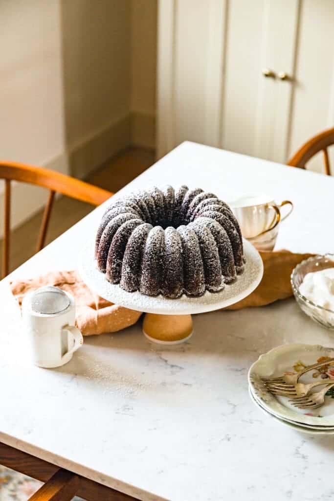 Picture of a chocolate bundt cake dusted in powdered sugar, displayed on a white porcelain cake plate atop a marble countertop. 