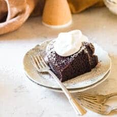 A slice of the perfect Chocolate bundt cake with a dollop of whipped cream atop a ceramic plate with forks resting nearby.