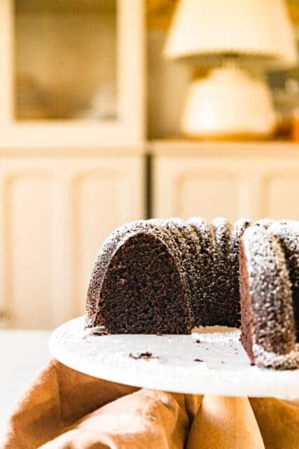 The Perfect Chocolate Bundt Cake, dusted with powder sugar displayed on a white cake plate missing a few slices.