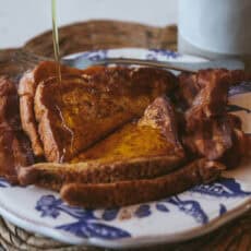 Golden cinnamon French toast slices topped with crispy bacon on a blue and white plate as warm maple syrup is poured over the toast.