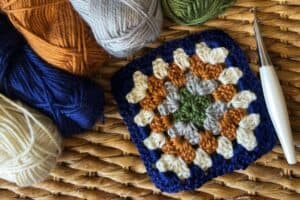 Colorful crochet granny square with yarns and crochet hook on woven surface.