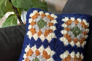 Crochet pillow featuring multicolored granny squares in blue, white, orange, and green.