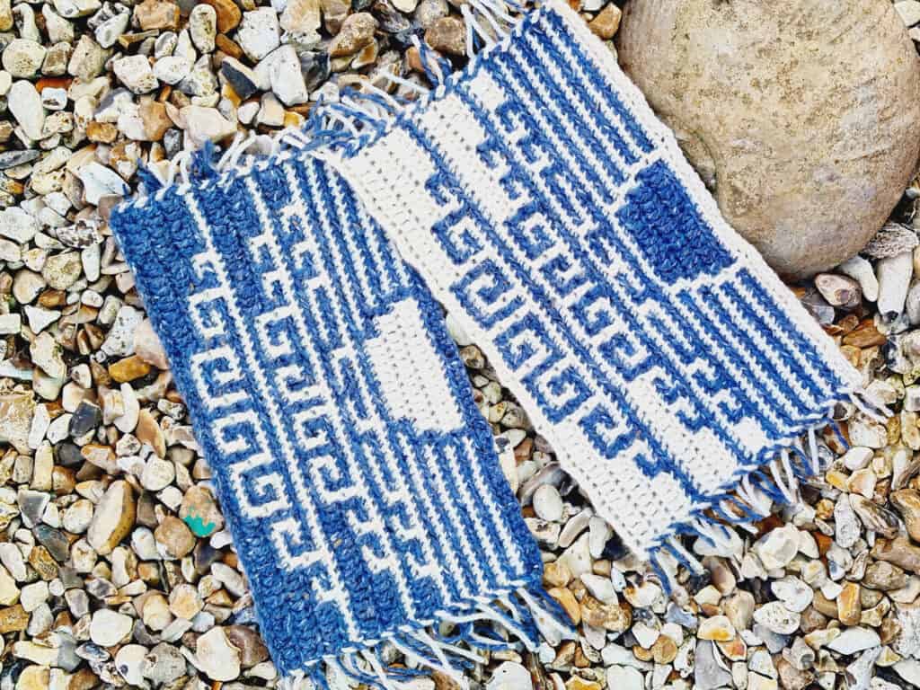 Two variations of a mosaic crochet design of a setting sun over ocean waves, made in with blue and white. Pieces are arranged on a pebble beach.